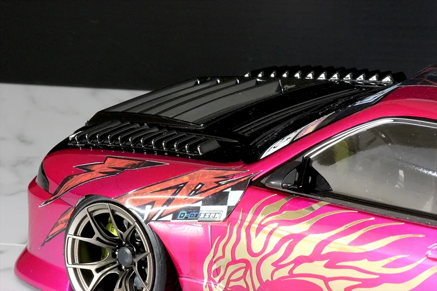 Close-up of an RC car body shell painted pink with black hood vents, decals, and custom wheels.
