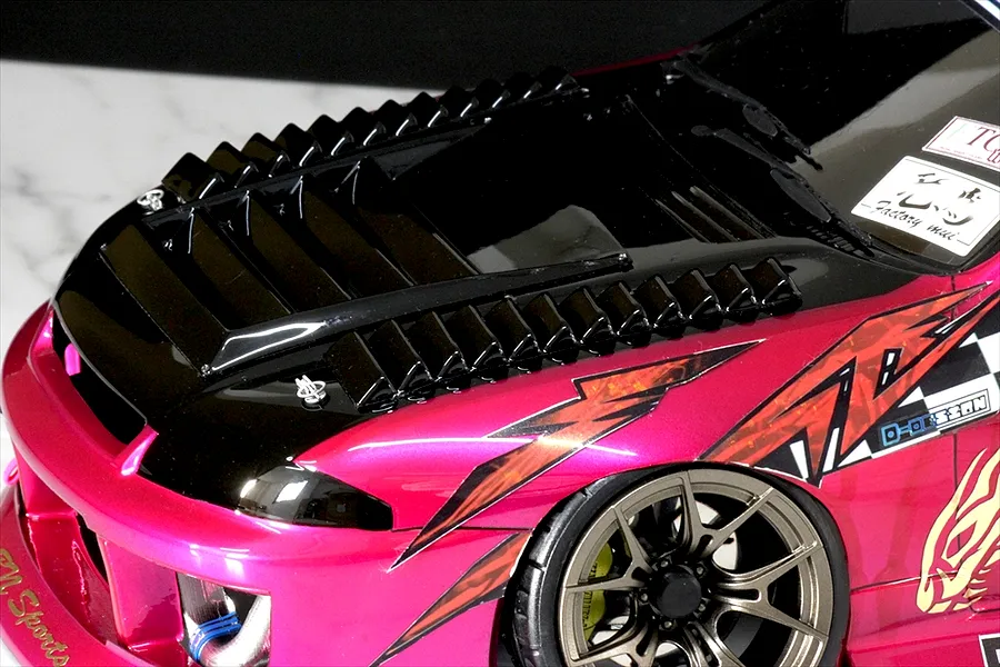 Close-up of an RC drift car body shell featuring black hood louvers and duct parts installed on a metallic pink body.