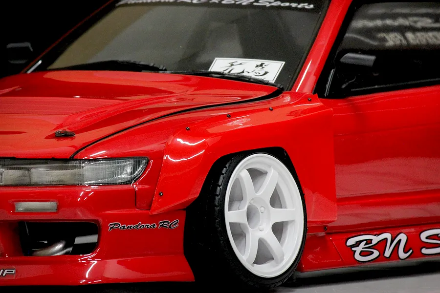Close-up of a red RC drift car body installed with wide over-fenders and white six-spoke wheels.
