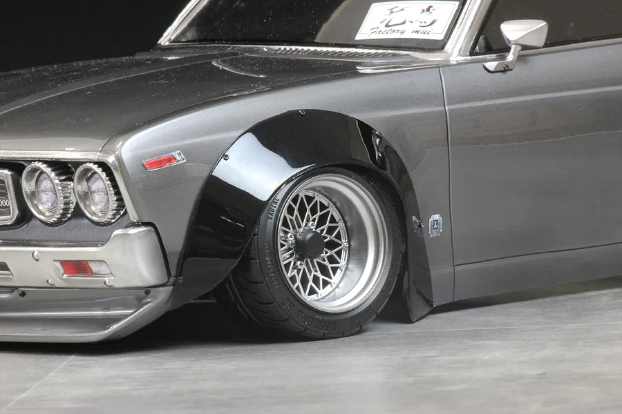 Close-up of a grey RC car body shell fitted with a black overfender set, silver mesh wheels, and low-profile tires.
