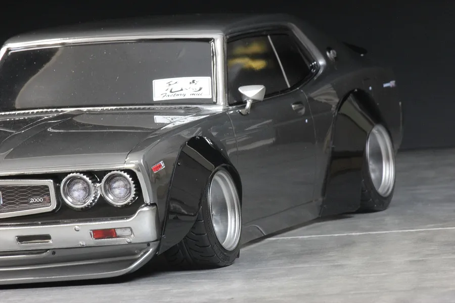 Close-up of a grey RC car body installed with wide black overfenders over deep-dish wheels against a dark background.