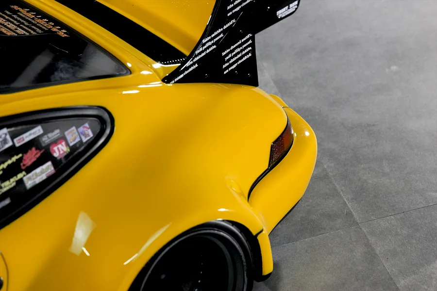 Close-up of the rear quarter, fender, and large wing of a yellow Pandora RC RWB 993 body shell on a grey floor.