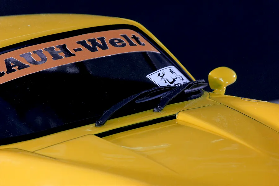 Close-up of a yellow RC car body shell showing the windshield, wipers, side mirror, and Rauh-Welt windshield banner.