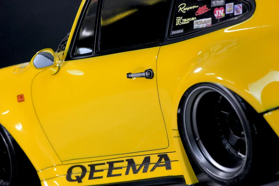 Close-up of the side profile of a yellow RC car body shell, featuring wide fender flares, black door handles, and sponsor decals.