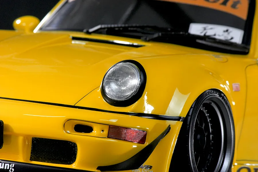 Close-up of the front left section of a yellow RWB 993 RC car body shell showing the headlight, bumper, and wheel.
