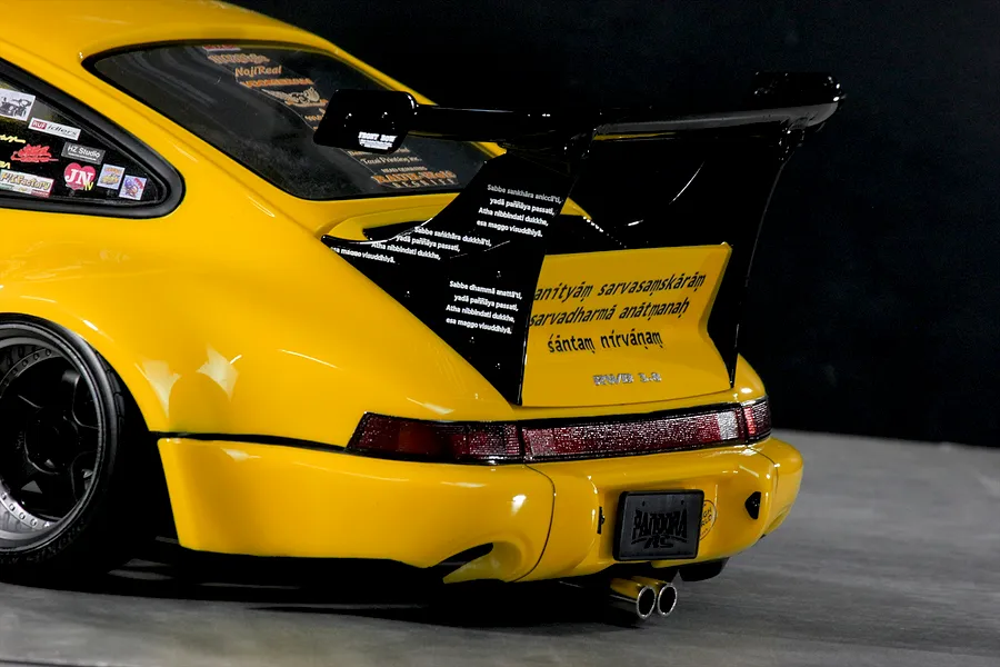 Close-up rear angle of a yellow RC body shell featuring a large black spoiler, custom decals, wide fenders, and twin exhaust pipes.