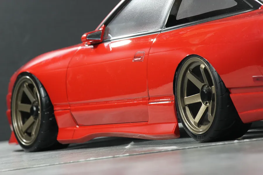 Close-up side profile of a red RC drift car body shell with bronze six-spoke wheels on a grey surface.