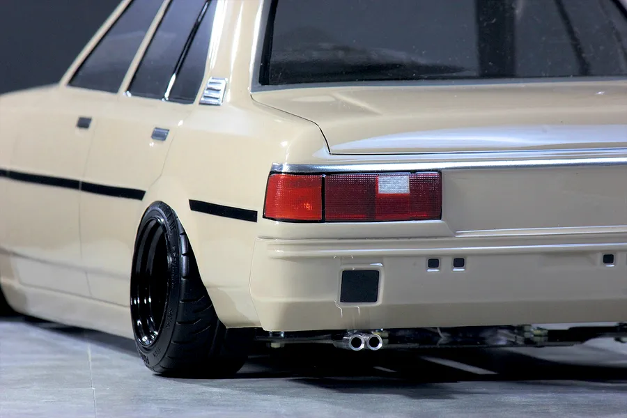 Close-up rear quarter view of a cream-colored Toyota Corolla KE70 RC drift car body showing dual exhaust pipes, deep dish wheels, and tail lights.