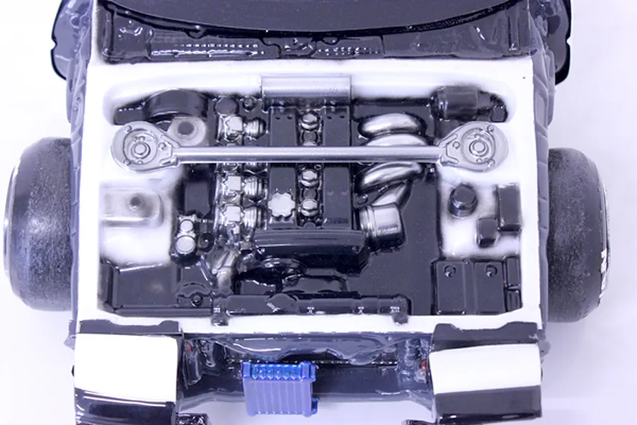 Top-down view of a detailed model car engine bay featuring a painted engine block, strut bar, and components inside a white chassis.