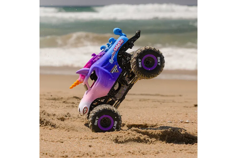 Losi Mini LMT Sparkle Smash RC monster truck performing a vertical wheelie on a sandy beach with ocean waves in the background.