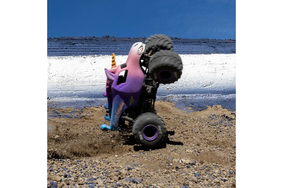 Losi Mini LMT Sparkle Smash RC monster truck performing a vertical stand on dirt track against a blue sky.
