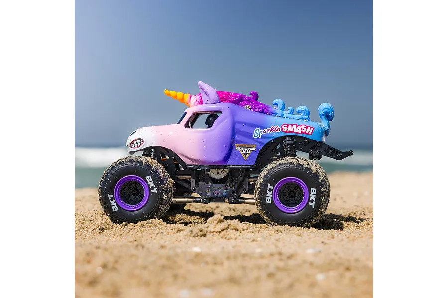 Losi Mini LMT Sparkle Smash RC Monster Truck driving on a sandy beach under a clear blue sky.