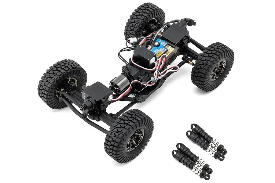 Top-down studio shot of a 1/24 scale RC rock crawler chassis without body shell, showing electronics, motor, wheels, and loose shocks on a white background.
