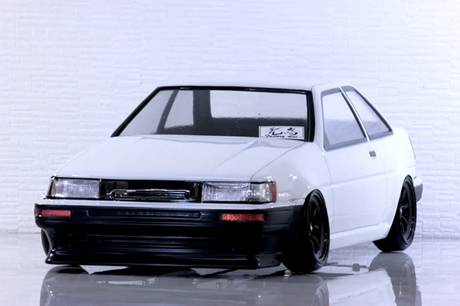Front three-quarter view of a white Toyota AE86 Corolla Levin RC drift car body with black wheels on a reflective surface.