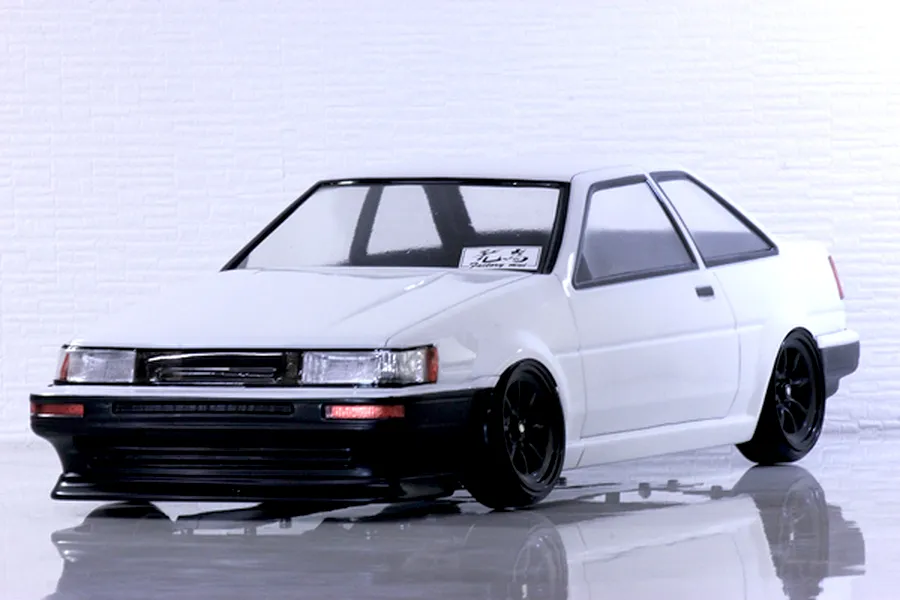 Studio photo of a white Toyota AE86 Corolla Levin 2-door RC car body with black wheels and front bumper against a light brick wall background.