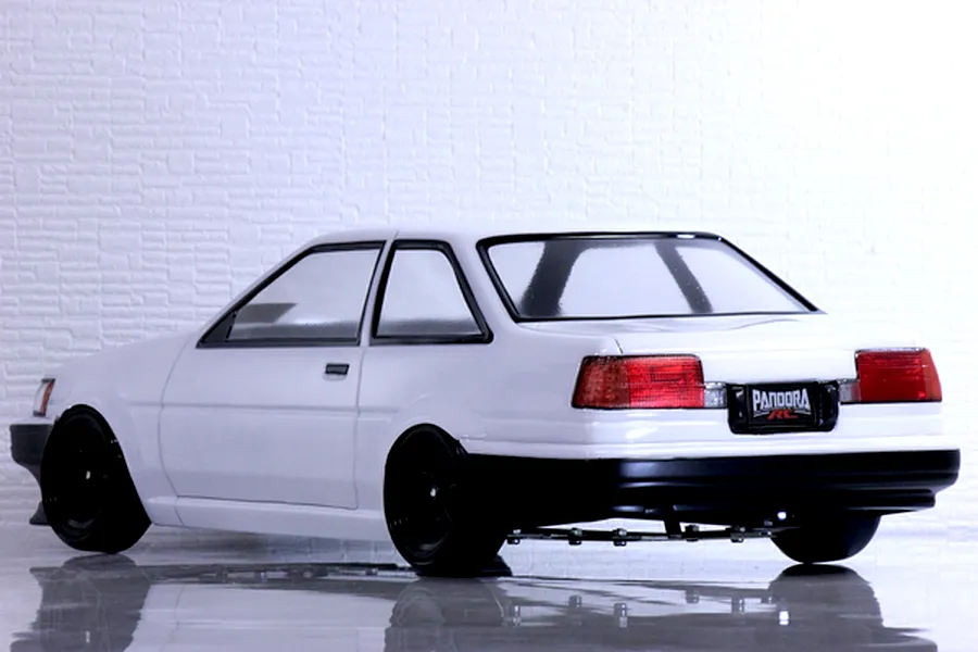 Rear-three-quarter view of a white Toyota AE86 Corolla Levin RC car body with black wheels on a reflective surface.