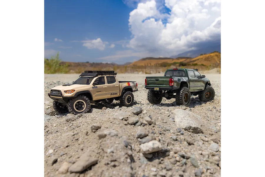 Two remote control Toyota Tacoma rock crawler trucks in beige and green parked on a rocky outdoor trail against a blue sky.