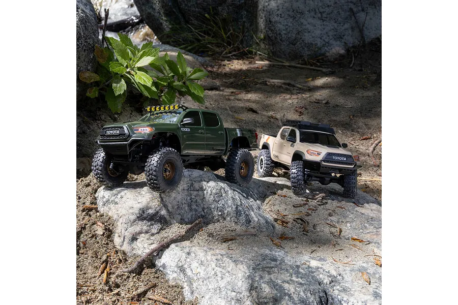 Two remote control Toyota Tacoma rock crawler trucks in green and tan driving outdoors on rocks and soil.