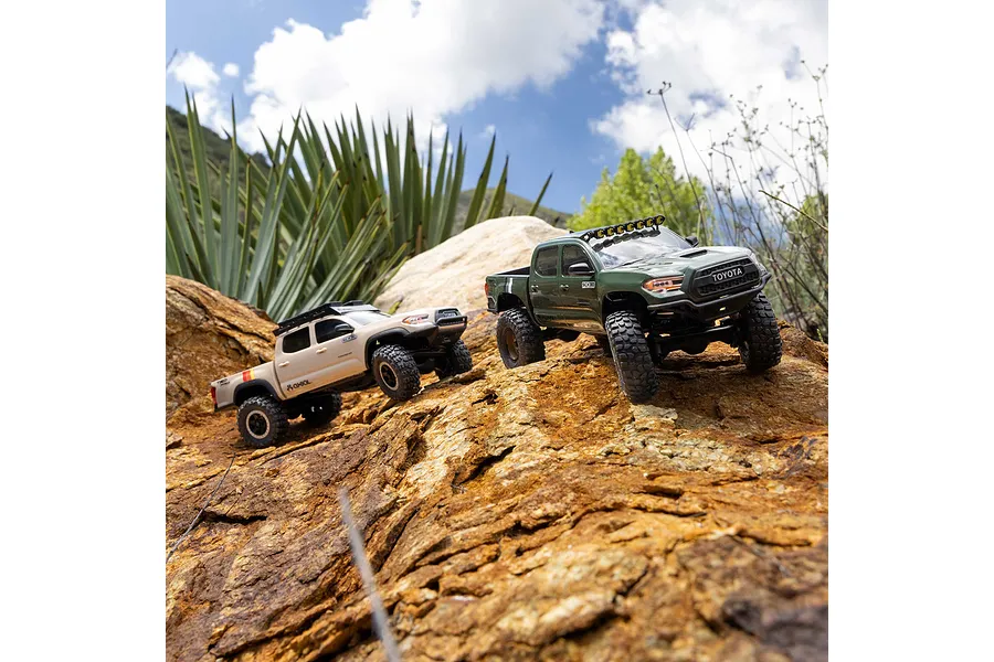 Two Axial Toyota Tacoma RC rock crawlers navigating rugged rocky terrain outdoors with green foliage and cloudy blue sky background.