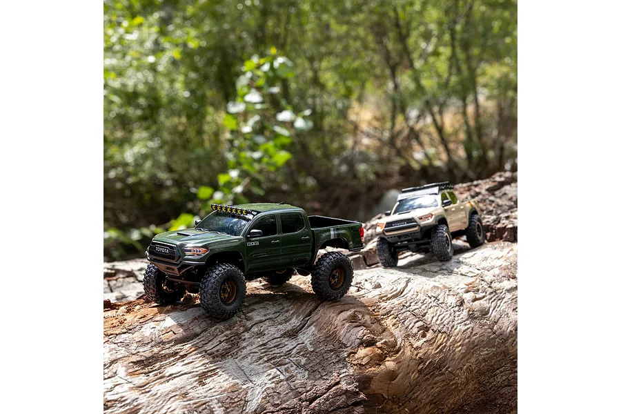 Two Axial RC crawler trucks driving along a large fallen tree trunk outdoors in a leafy forest.