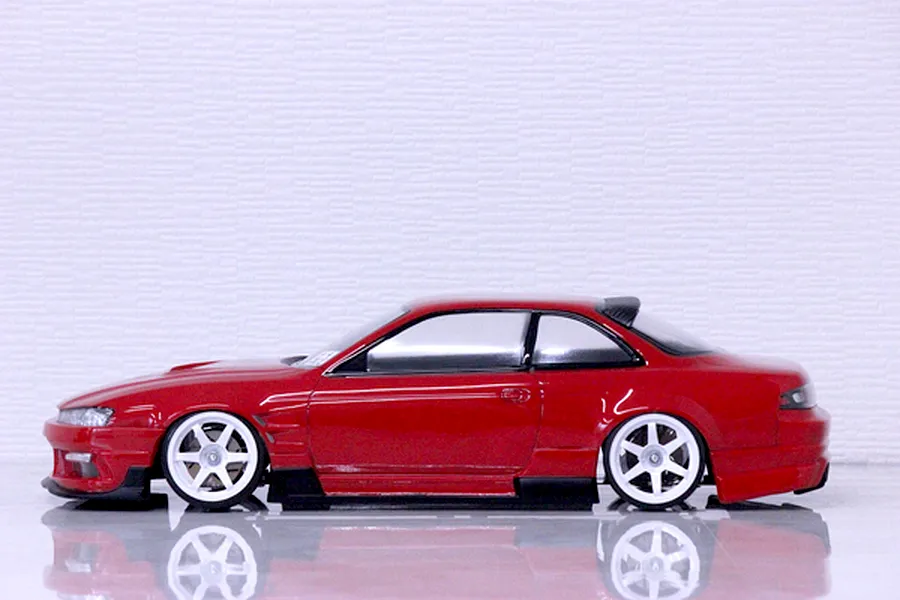 Side profile view of a red RC drift car body on a reflective surface against a white textured background.