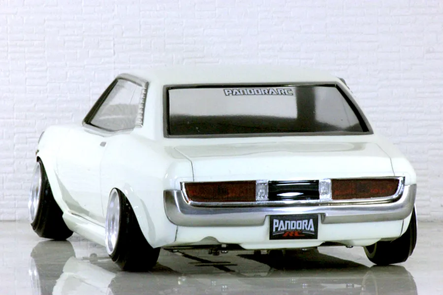 Rear three-quarter view of a white Pandora RC Toyota Celica 1600GT drift car body shell on a reflective surface with a brick wall backdrop.