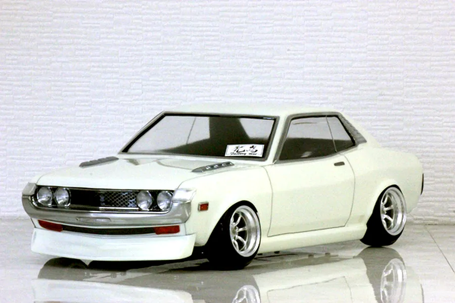 White Toyota Celica 1600GT RC drift car body shell mounted on a chassis with custom wheels and a textured white brick background.