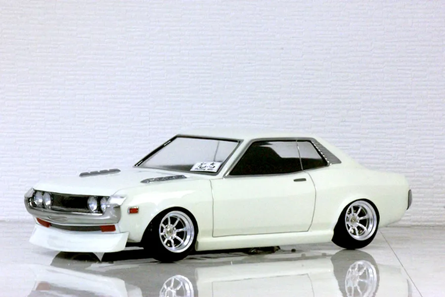 White Toyota Celica 1600GT RC body shell mounted on a chassis with custom wheels against a textured white brick background.