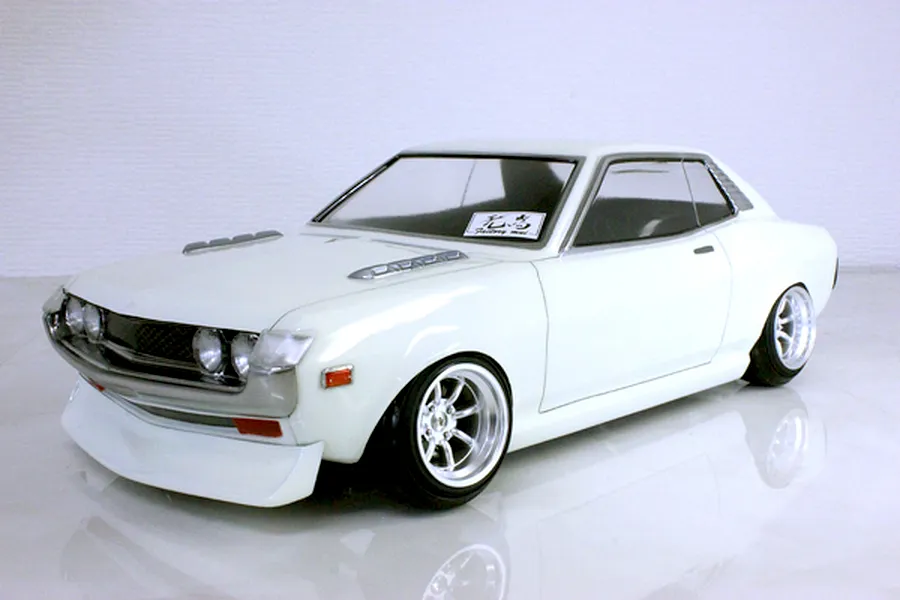 Front three-quarter view of a white Toyota Celica 1600GT RC body shell mounted on a chassis with custom wheels against a white background.