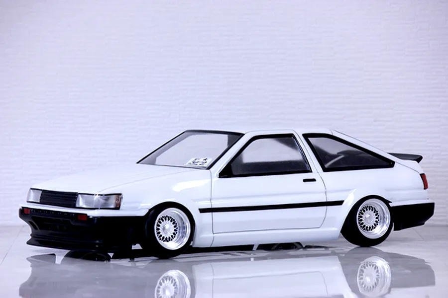 A painted white Toyota AE86 Corolla Levin 3-door RC body shell mounted on a chassis with wire-mesh wheels, sitting on a reflective surface.