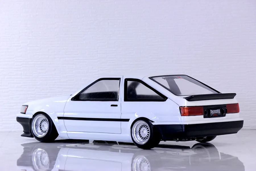 Side-rear profile view of a painted white Toyota AE86 Corolla Levin RC drift car body shell with black bumpers and mesh wheels.