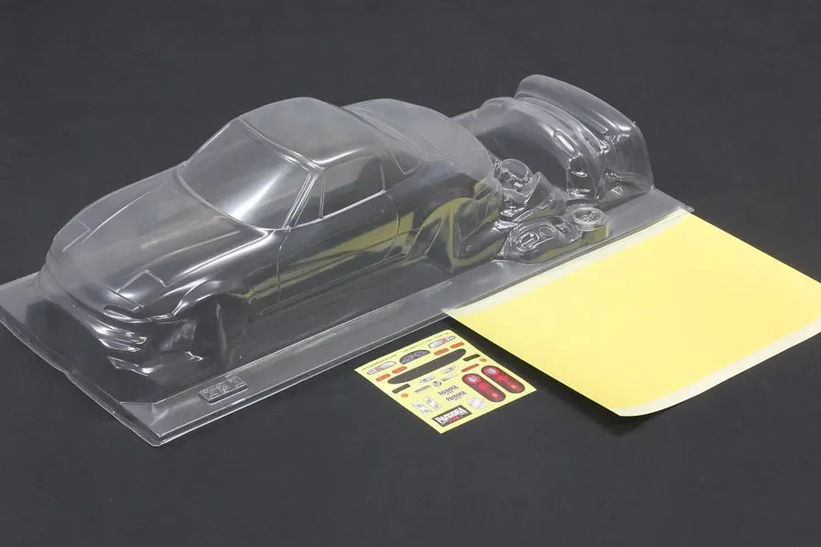 Unpainted clear polycarbonate RC car body shell with a mask sheet and sticker sheet on a dark surface.