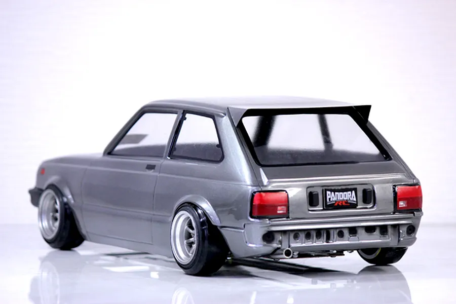 Rear-three-quarter view of a painted grey Toyota Starlet KP61 RC drift car body shell with Pandora RC license plate and custom wheels on a reflective white surface.