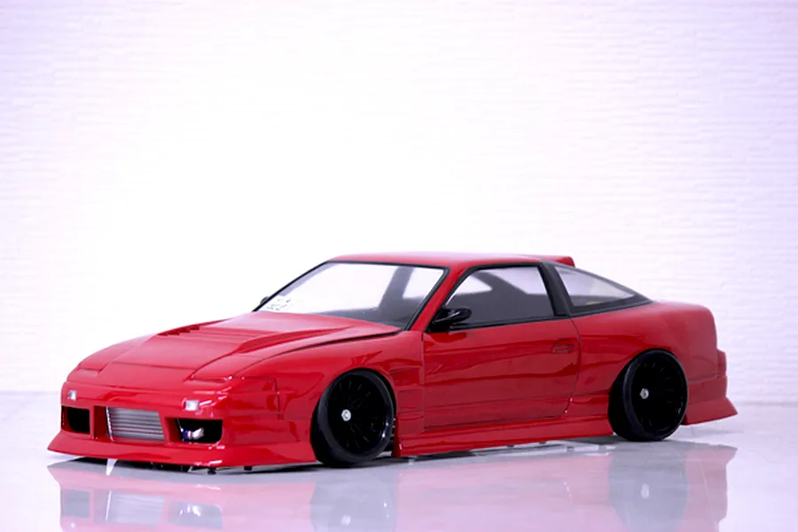 Red RC drift car body shell mounted on a chassis with black multi-spoke wheels on a light surface against a white textured background.