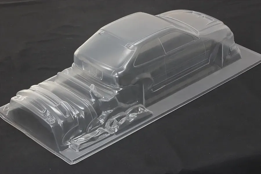 Unpainted clear polycarbonate RC car body shell on a dark background showing molded hatch and front bumper parts.