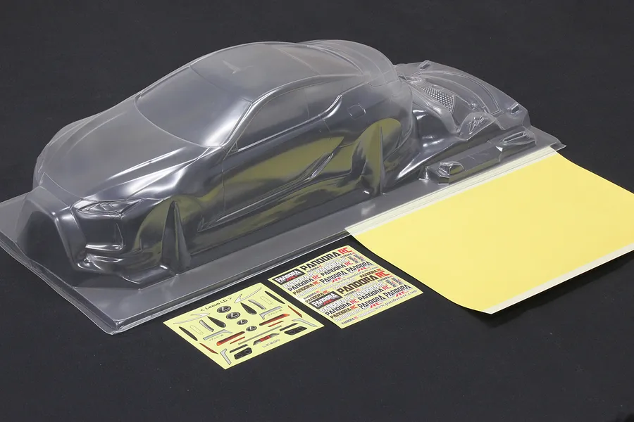 Unpainted clear polycarbonate RC car body shell with window masks and decal sheets on a dark surface.