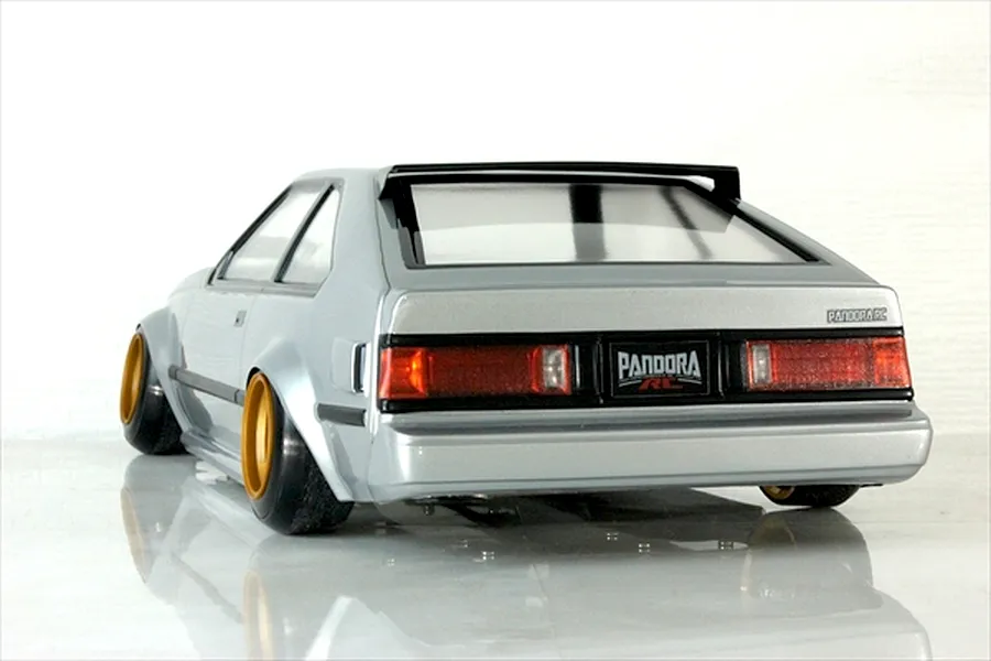 Rear-three-quarter view of a silver Toyota Celica RC drift car body shell with gold wheels and tinted windows on a reflective white surface.