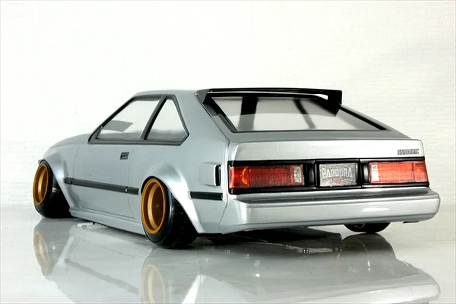 Rear three-quarter view of a painted silver RC drift car body on a reflective white surface with gold wheels and black trim.