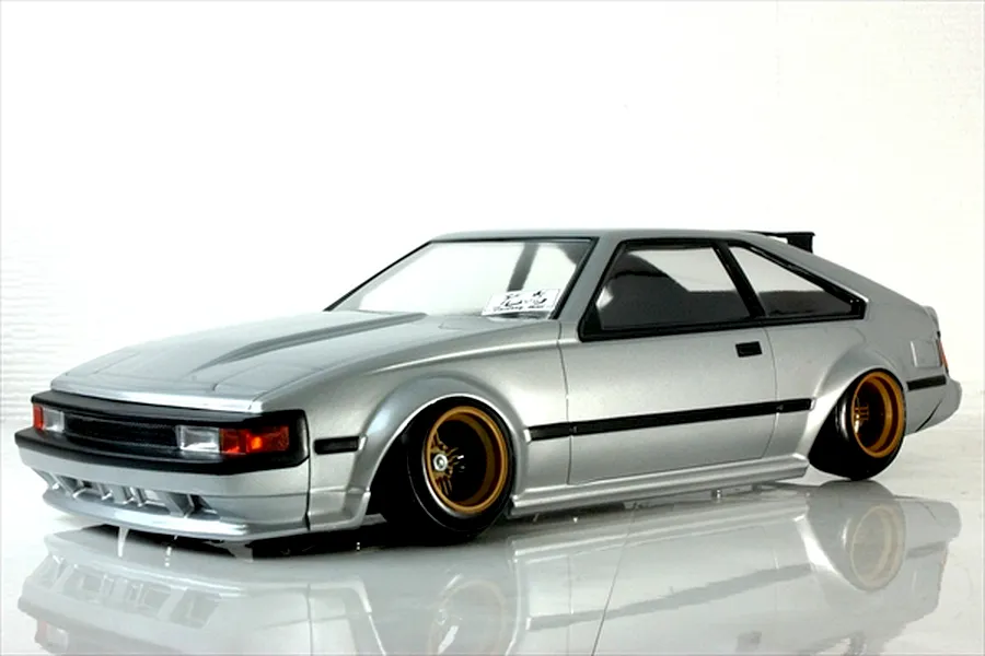 Silver Toyota Celica XX A60 RC drift car body shell mounted on a chassis with gold deep dish wheels against a white background.