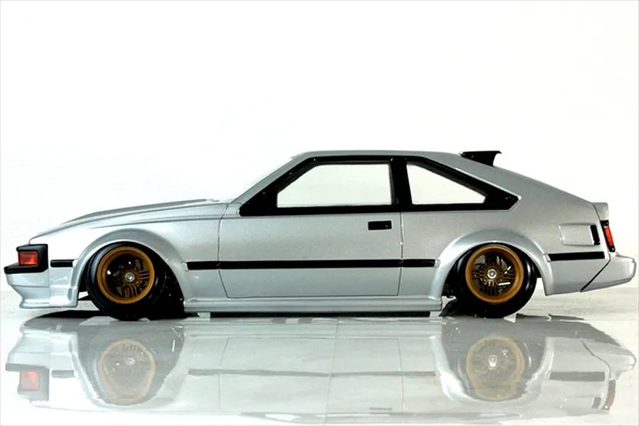 Side profile view of a silver Toyota Celica RC drift car body with gold mesh wheels on a white reflective surface.