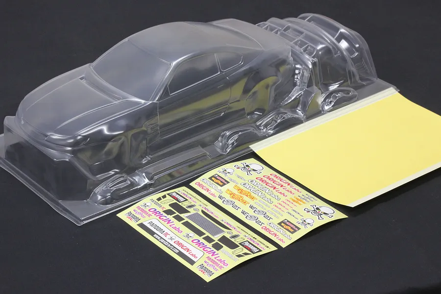 Clear unpainted RC car body shell with separate parts, decal sheet, and window mask sheet on a dark background.