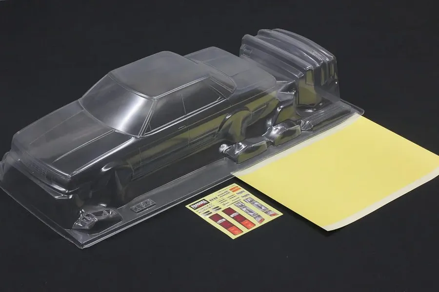 Unpainted clear polycarbonate RC car body shell with a sticker sheet and masking sheet on a dark background.