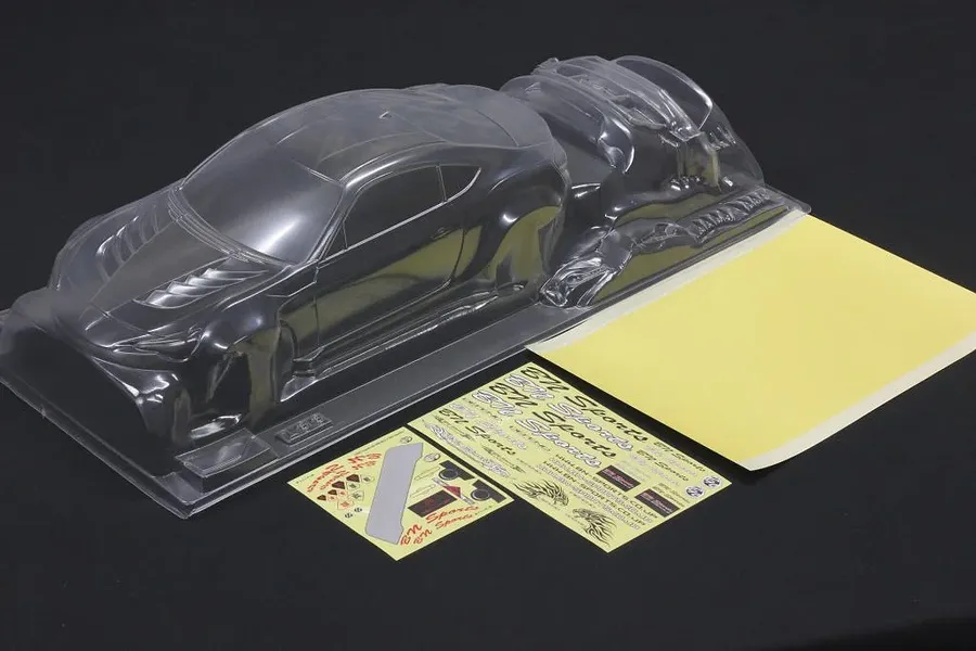 Unpainted clear RC car body shell with accessory parts and sticker sheets on a black surface