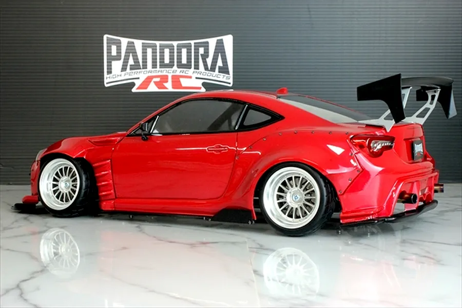 Side view of a red Pandora RC Toyota 86 body shell with wide body kit, large rear wing, and custom wheels on a reflective surface.