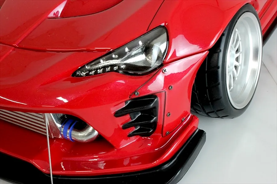 Close-up of the front left section of a red RC car body, showing the headlight, front wheel, bumper, and intercooler.