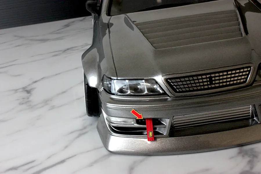Close-up front view of a grey RC drift car body showing the bumper, grille, and a red tow strap with an arrow pointing to it.