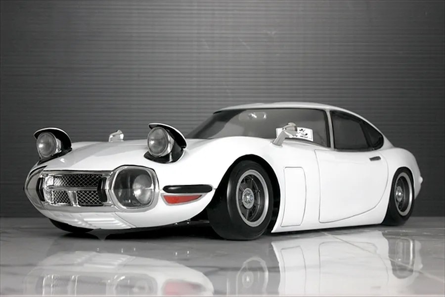 A front three-quarter view of a white Toyota 2000GT RC body shell with pop-up headlights open, parked on a reflective surface.