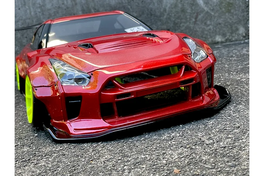 Low angle front view of a red RC drift car body featuring a wide body kit and bright fluorescent yellow wheels parked on asphalt.