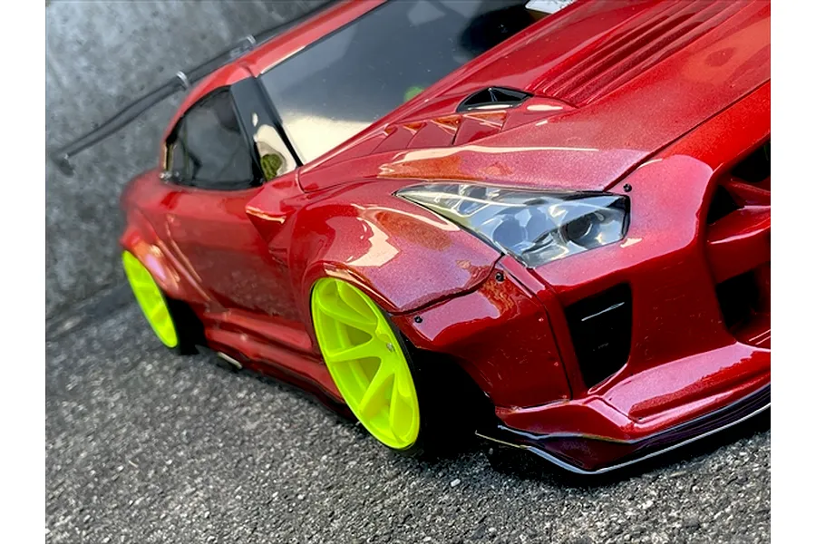 Close-up angled view of a red RC drift car body with wide fender flares and bright yellow wheels parked on asphalt.