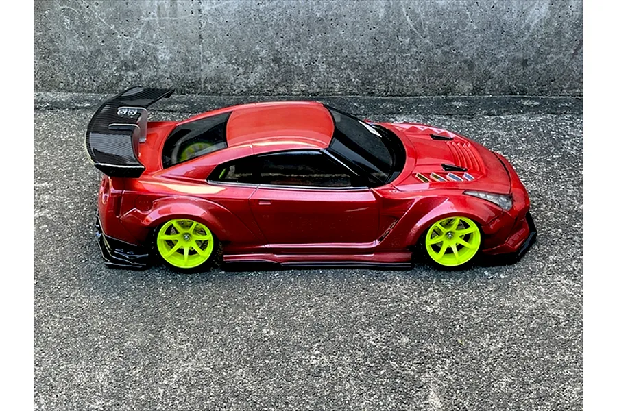 Side profile of a red metallic RC drift car body shell with a large rear wing and bright green wheels on asphalt.